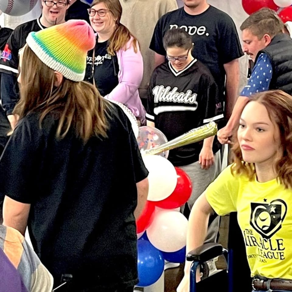 Diverse group of people, including a person in a rainbow hat, gathered with red, white, and blue balloons.